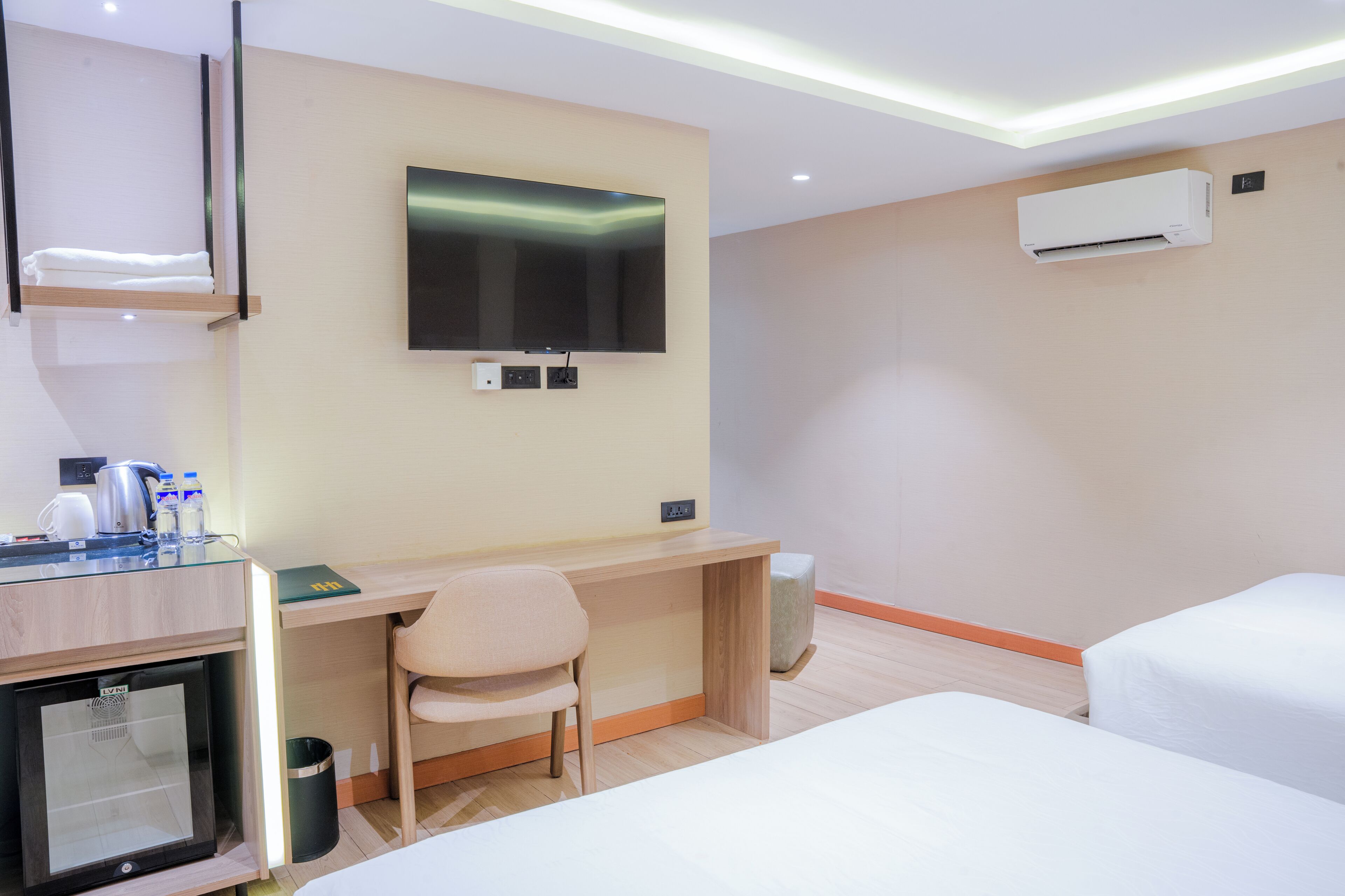 Ein Hotelzimmer mit Flachbildfernseher, Klimaanlage, Schreibtisch mit Stuhl, Minikühlschrank und einem Bett.