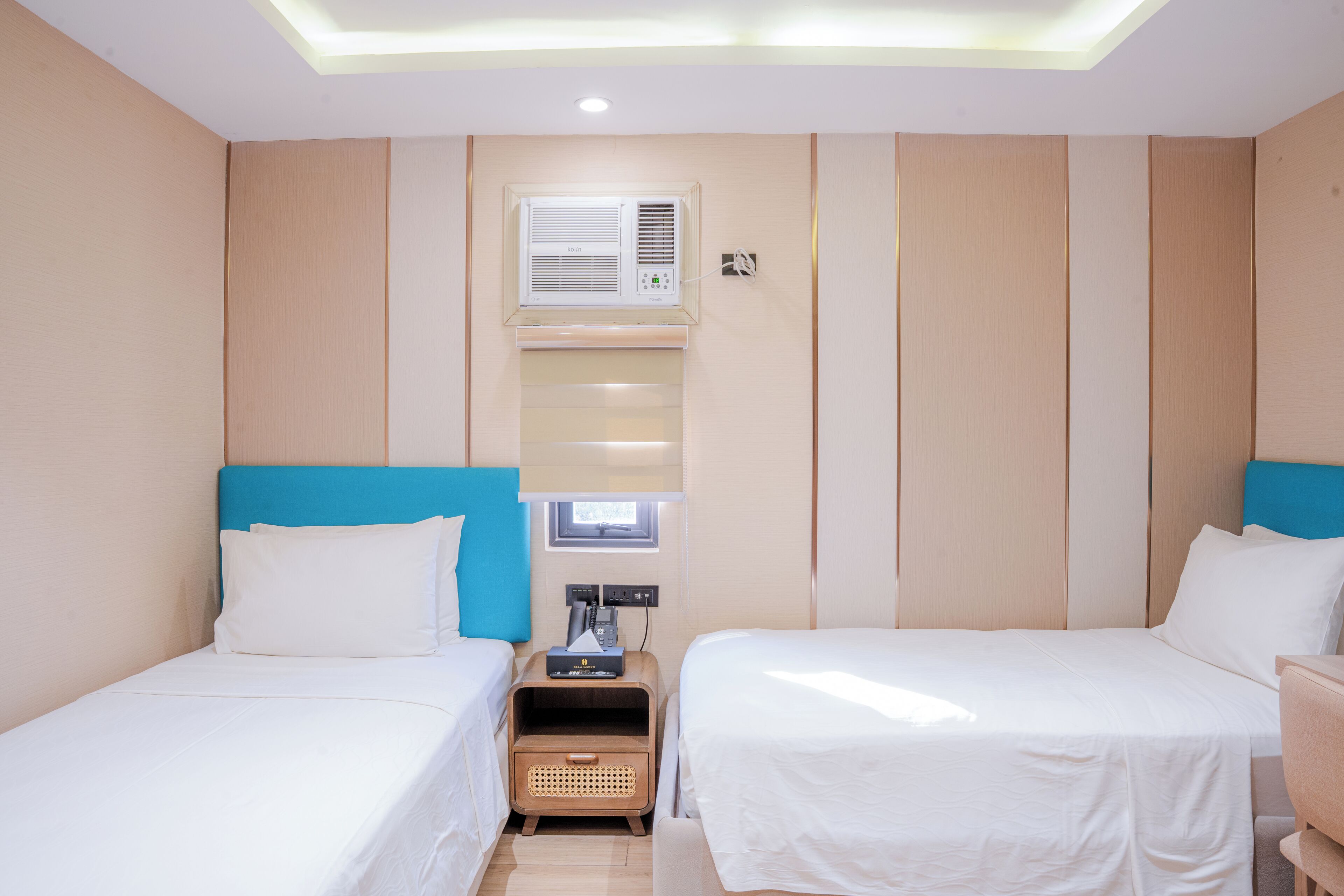 Habitación de hotel con dos camas, una lámpara del techo, un aire acondicionado y una mesita de noche con teléfono y lámpara.
