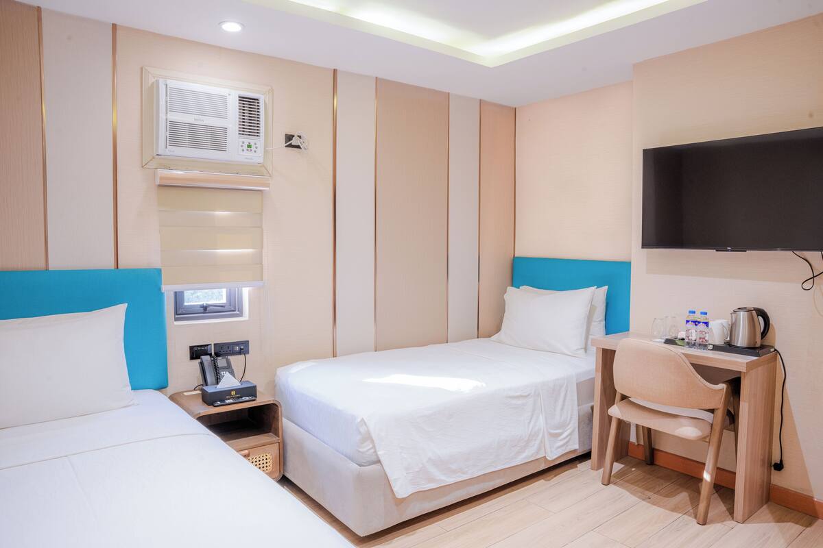 A hotel room with two beds, a desk with a chair, a flat-screen TV, an air conditioning unit, and a small window.