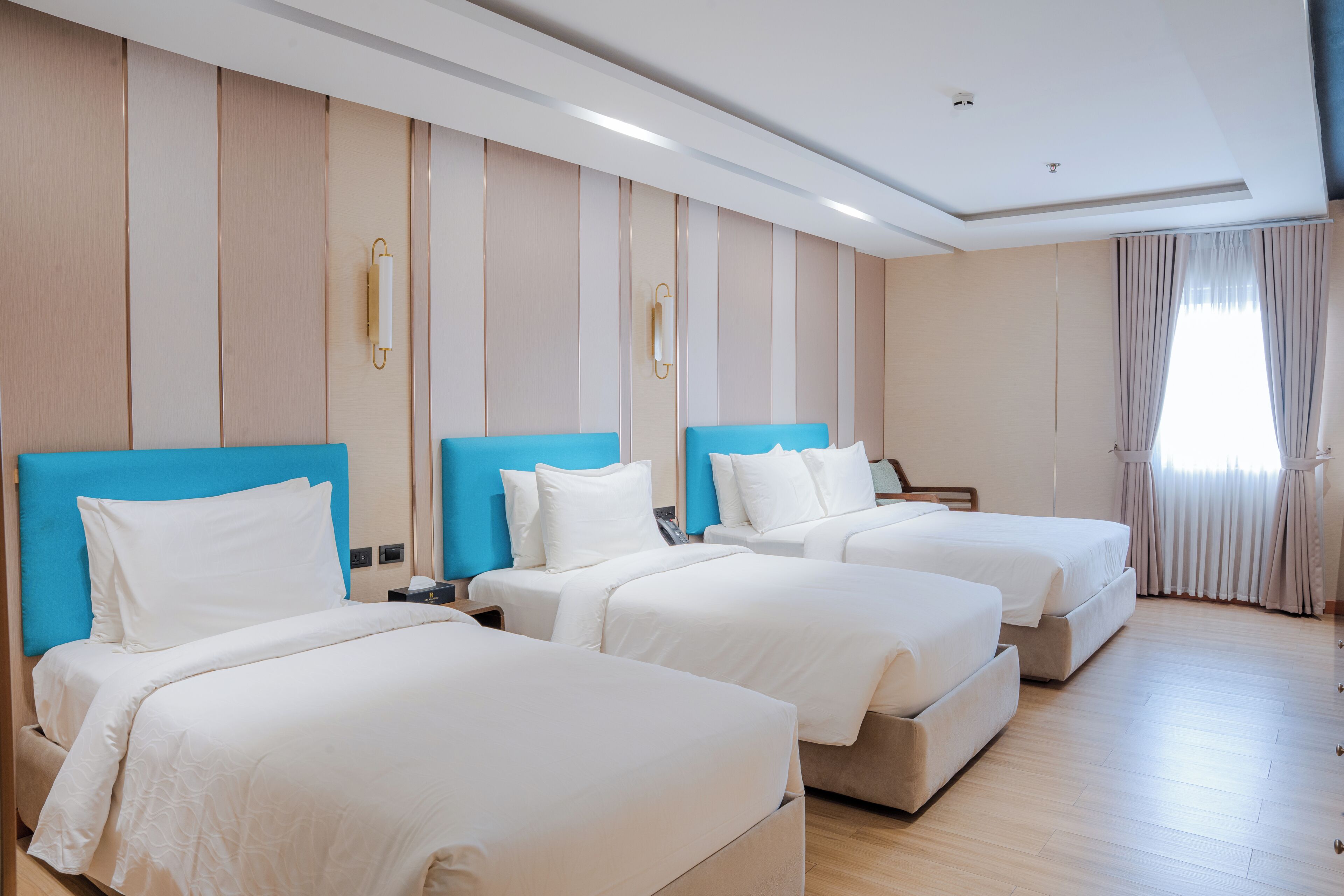 A hotel room with three beds, a wooden floor, and a large window with curtains.