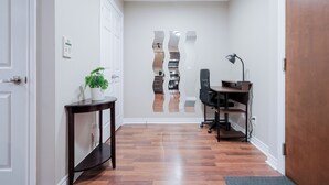 Un pasillo con suelo de madera, un escritorio con silla, una planta en maceta y un panel de seguridad en la pared.