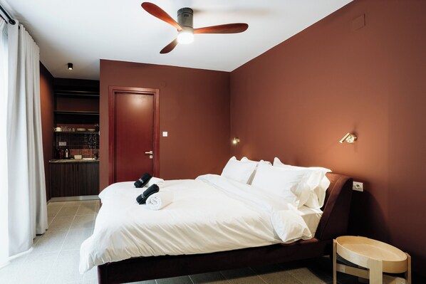 A hotel room with a bed, a ceiling fan, a small wooden chair, and a kitchenette in the background. - Canvas Color Your Stay (Volos)
