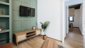 Una sala de estar moderna con un televisor de pantalla plana montado en una pared verde, una consola de madera con una planta, y un área de comedor visible a través de un vano abierto.