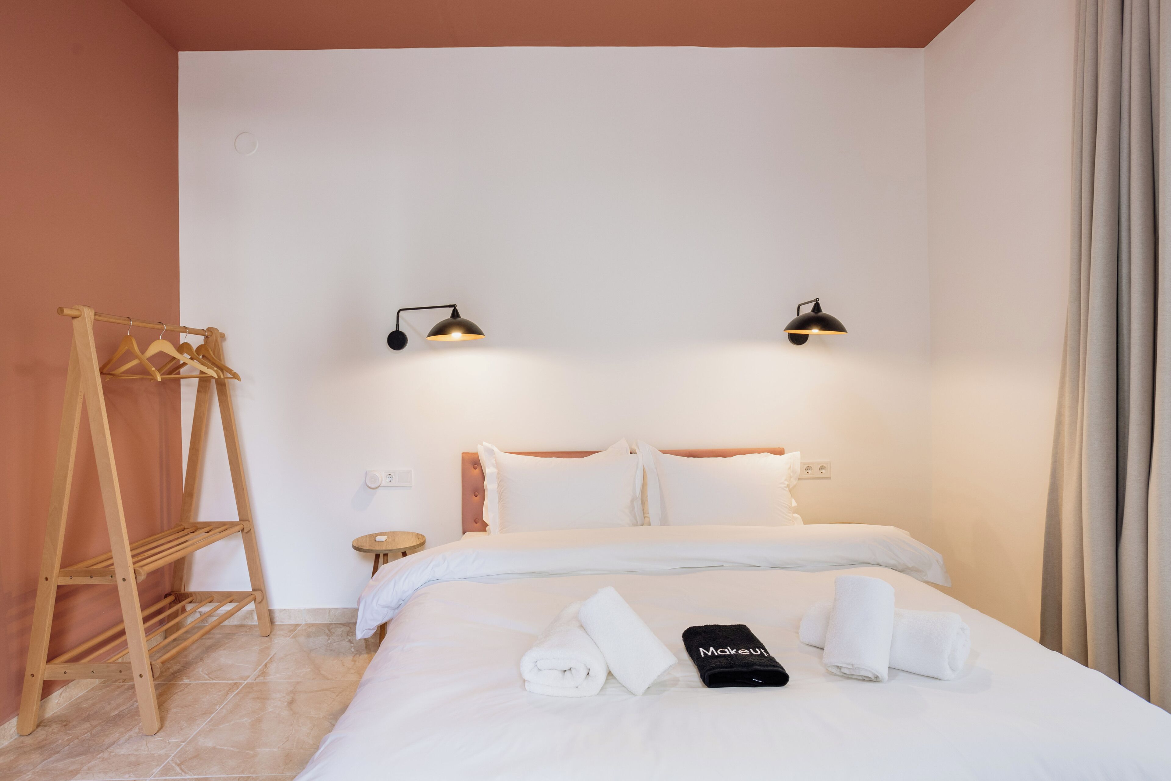 Ein Hotelzimmer mit einem Bett, zwei Nachttischlampen, einem Holzkleiderhaken und einem ordentlich bezogenen Bett mit Handtüchern und einem schwarzen Gegenstand darauf.