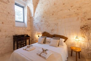Ein Schlafzimmer mit rustikaler Steinwand, einem Holzkopfteil, einem Bett mit weißer Bettwäsche, einem kleinen Holztisch und einem Nachttisch mit Lampe.