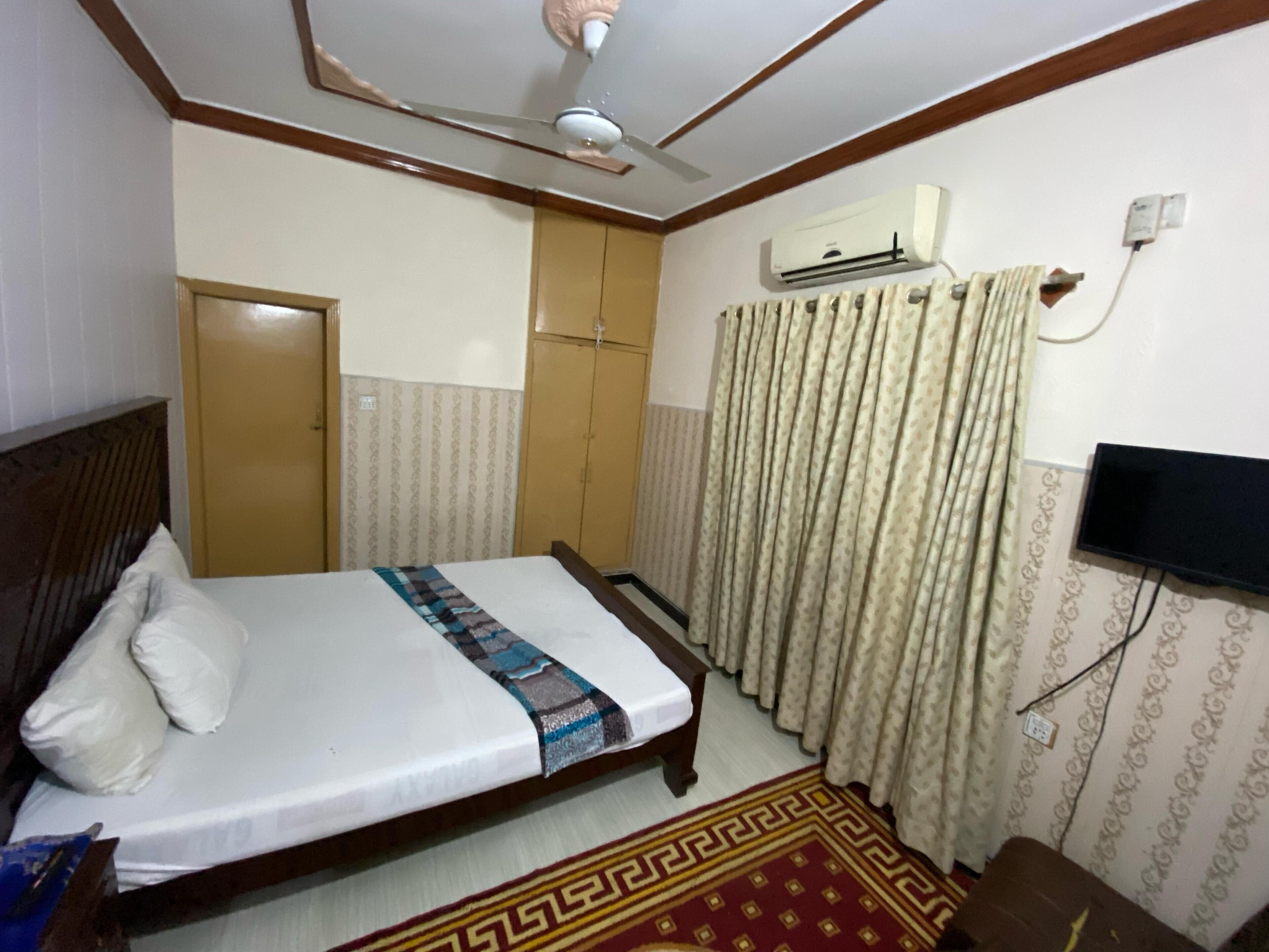 A hotel room with a bed, a ceiling fan, an air conditioning unit, a television, a wardrobe, and a patterned curtain.