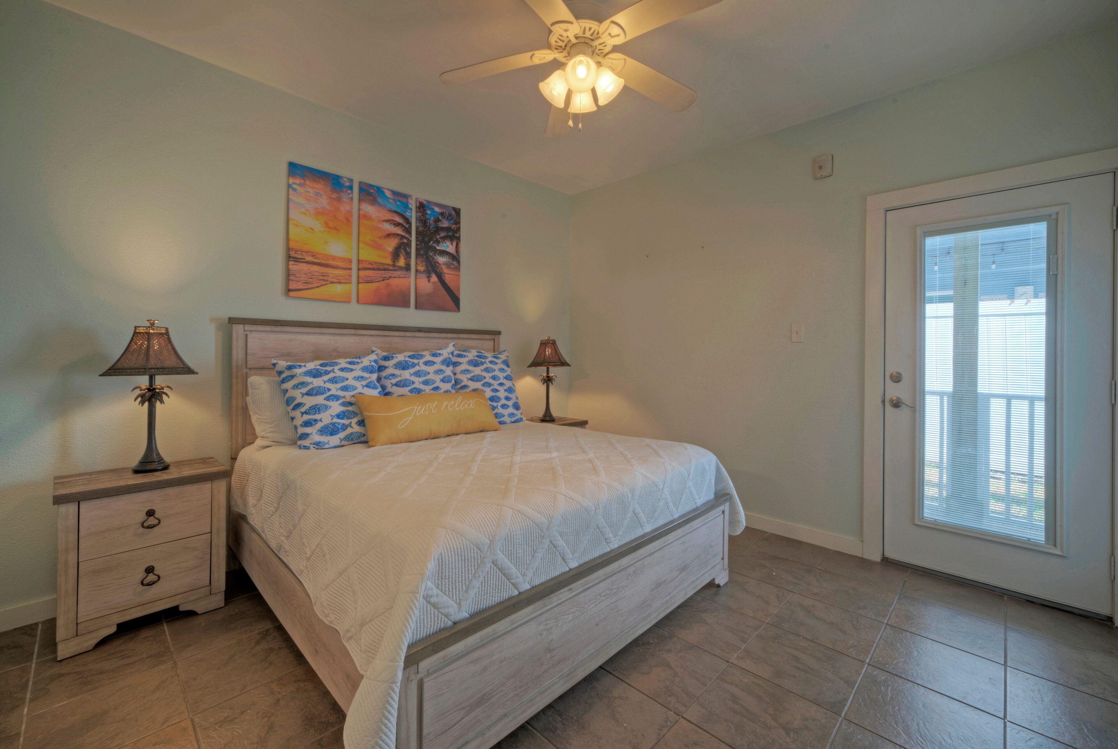 A bedroom with a bed, two nightstands, a ceiling fan, a door with a view, and a sunset painting on the wall.