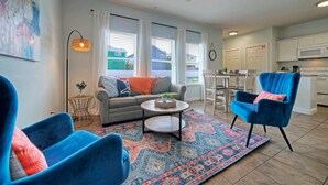A living room with a sofa, two armchairs, a coffee table, and a patterned rug. There is a dining area with a table and chairs, and a kitchen with cabinets and appliances.
