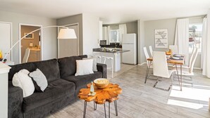 A modern living room with a dark grey sofa, white cushions, a wooden coffee table, and a dining area with white chairs and a table. - 7 Bungalows | Steps to Pier + Beach | Ocean Views, Outdoor Bar, Firepit, Lounge (Cayucos)