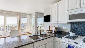 A modern kitchen with white cabinets, a granite countertop, a stove, and a large window offering a view of the sea. - 7 Bungalows | Steps to Pier + Beach | Ocean Views, Outdoor Bar, Firepit, Lounge (Cayucos)