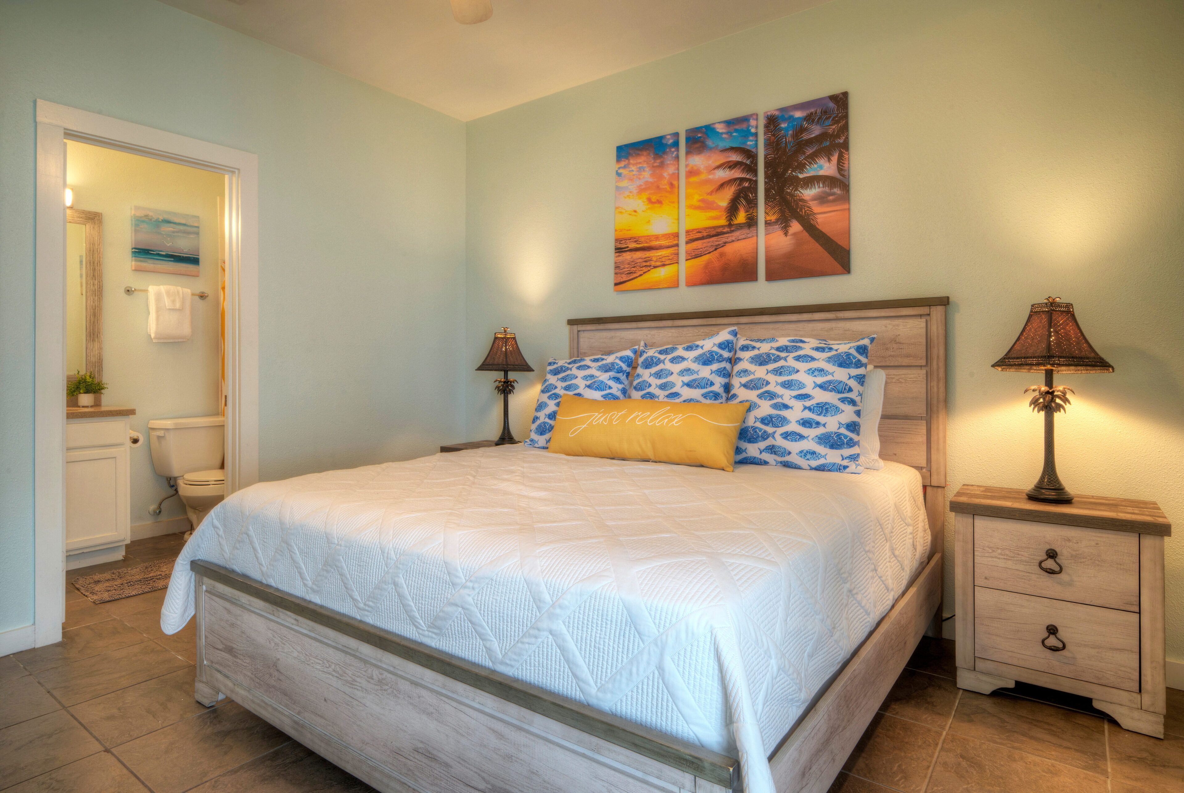 A bedroom with a bed, a nightstand, a lamp, a painting, and a bathroom visible through an open door.