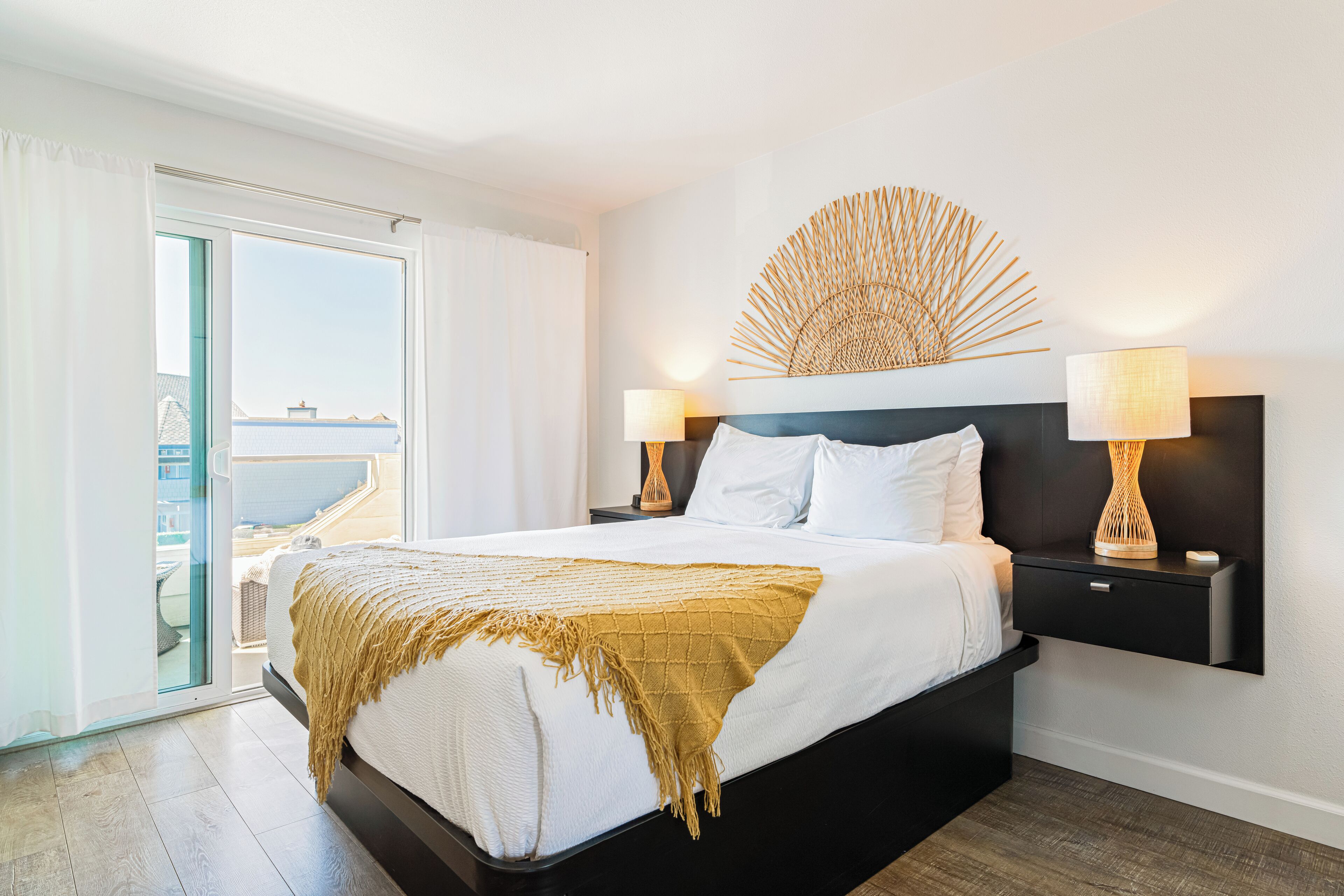 Quarto de hotel moderno com uma cama grande, uma mesa de cabeceira, uma luminária na parede e vista para o mar por uma porta de correr.