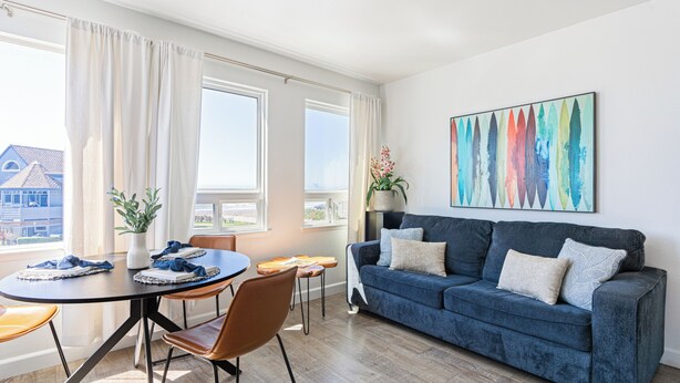A modern living room with a blue sofa, a dining table with chairs, and a large window with curtains. - 2 Cayucan Bungalows | Steps to Beach, Pier | Shared Outdoor Bar, Firepit, Lounge (Cayucos)