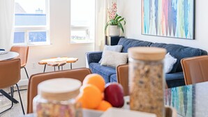 Ein modernes Wohnzimmer mit einem blauen Sofa, einem Esstisch mit Stühlen, einer Topfpflanze und einem farbenfrohen, abstrakten Gemälde an der Wand.