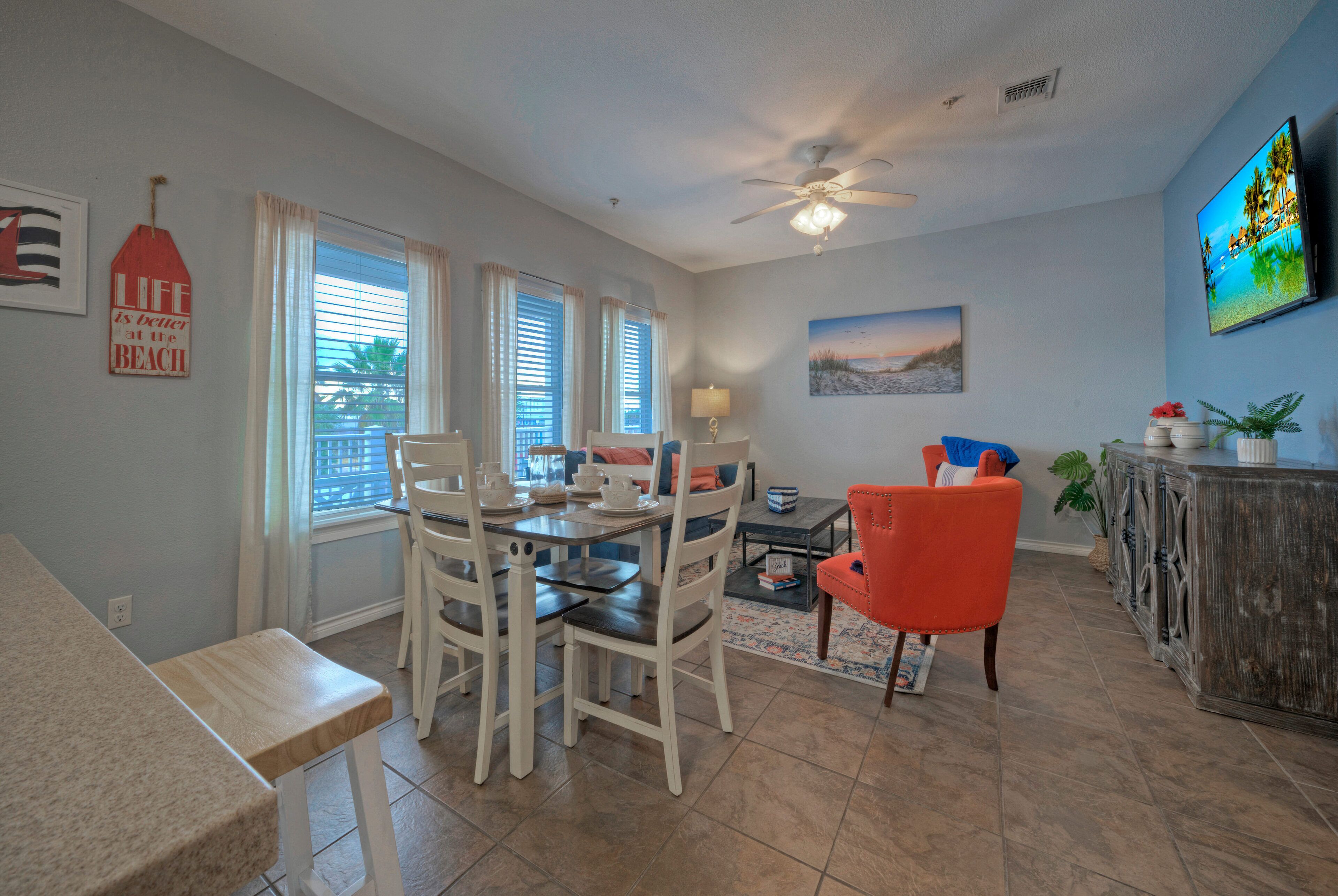 A dining area with a table set for four, white chairs, a ceiling fan, and a view of the outdoors through large windows.