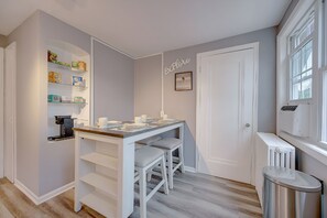 Une petite cuisine avec un plan de travail blanc, deux tabourets, une machine à café et une étagère remplie de produits alimentaires.