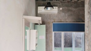 A room with a bed, a window with a view, a wall-mounted air conditioner, and a door leading to another room. - Rumah Betawi Kalipasir (Jakarta)