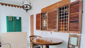 A room with a round wooden table, two chairs, a stool, and a metal bench. There are two wooden windows with shutters, a green door, and a ceiling fan. - Rumah Betawi Kalipasir (Jakarta)