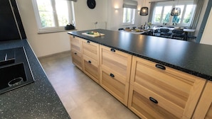 A kitchen with a large island, wooden cabinets, and a black countertop. There are windows with curtains, a clock on the wall, and a dining area visible in the background.
