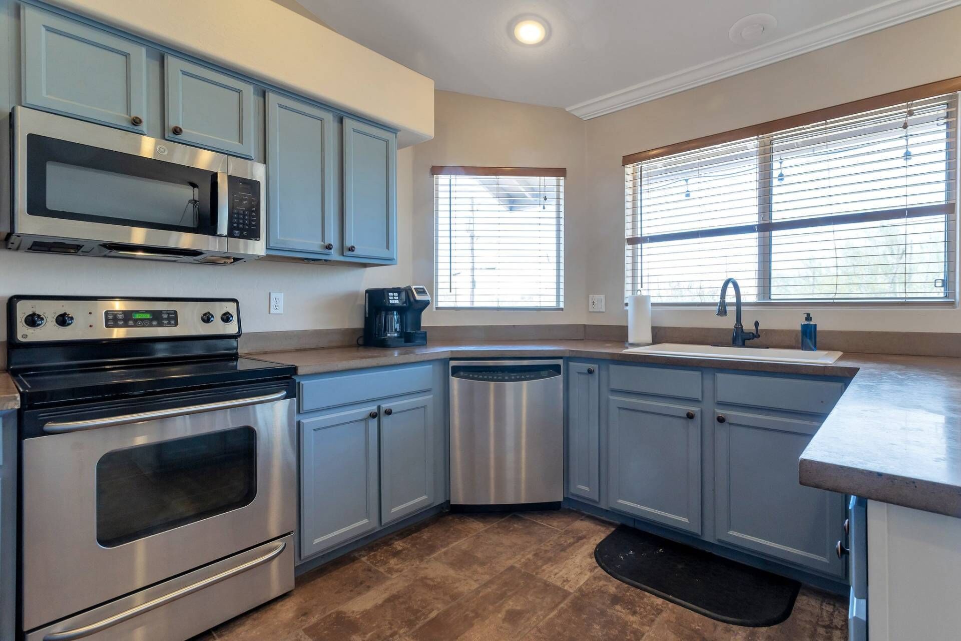 A kitchen with stainless steel appliances, light blue cabinets, a dishwasher, and a window with blinds.