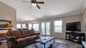 A spacious living room with a brown leather couch, a wooden coffee table, a colorful rug, a ceiling fan, a television, and a dining area in the background.