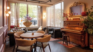 A cozy living room with a piano, leather sofas, a dining table, and a large window with curtains. - Boutique Hotel de Statie (Valkenswaard)