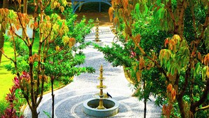 Um caminho circular de tijolos leva a uma fonte central, cercada por vegetação exuberante e uma pérgola azul ao fundo.