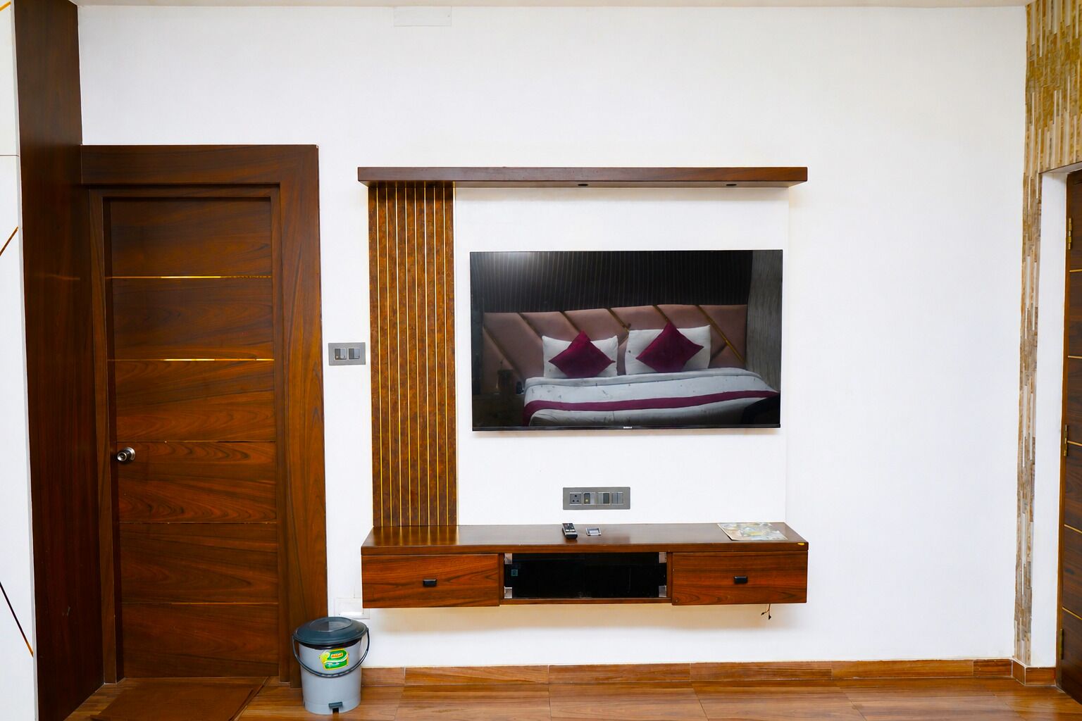 A bedroom with a bed, a wooden door, a small trash bin, and a wall-mounted shelf.