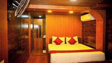 Una habitación de hotel con techo de madera, una cama con ropa de cama blanca y roja, un ventilador de pie y un aire acondicionado instalado en la pared.