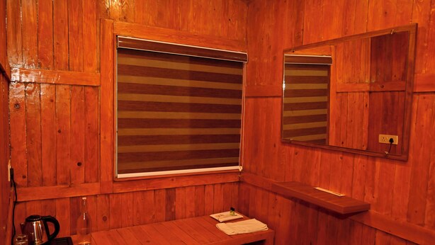 A wooden sauna room with a bench, a window with blinds, a mirror, and a small table with a kettle and cups. - WINNERS HILL RESORTS (Coimbatore)