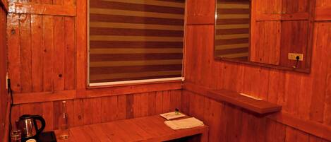 A wooden sauna room with a bench, a window with blinds, a mirror, and a small table with a kettle and cups.
