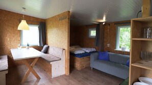 Ett litet rum med trägolv, en säng med en blå madrass, en grå soffa, ett litet bord med en lampa och en hylla med tallrikar.