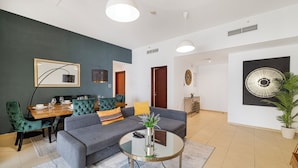 A modern living room with a grey sofa, a round coffee table, a dining area with a wooden table and green upholstered chairs, and a large abstract painting on the wall. - Elegant 1Br Apart With Beach Access In Dubai Creek (Dubai)