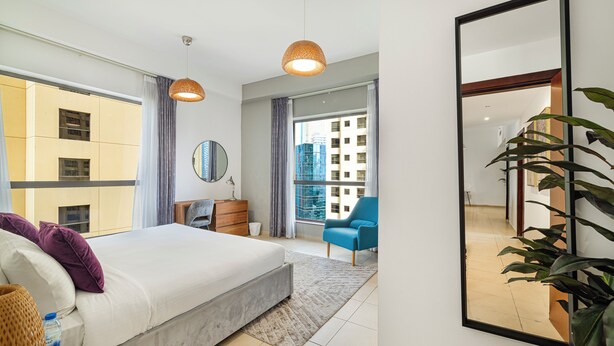 A modern bedroom with a large bed, a dresser, a chair, and a large window offering a city view. - Elegant 1Br Apart With Beach Access In Dubai Creek (Dubai)