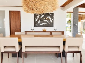 A modern dining area with a large wooden table, beige upholstered chairs, a hanging light fixture, and a black and white abstract painting on the wall. - Villa Takali (Pacific Harbour)