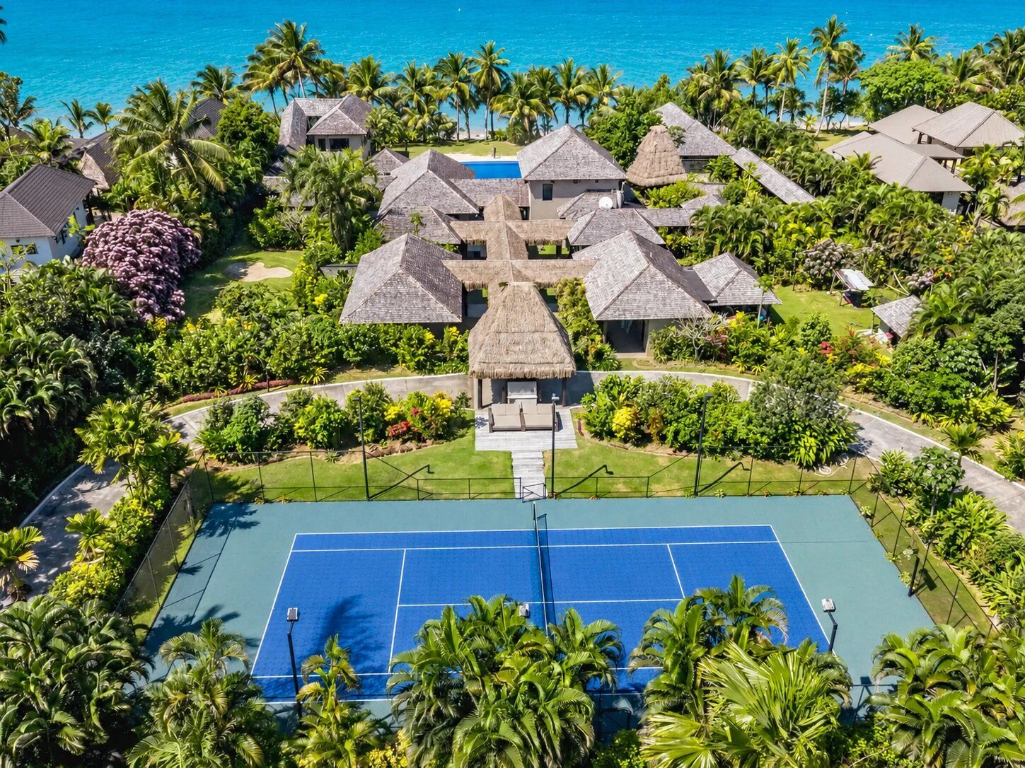 Un complexe hôtelier comprenant plusieurs bâtiments à toits de chaume, un court de tennis et une vue sur l’océan.