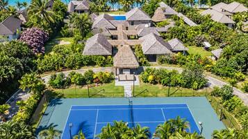 Un complexe hôtelier comprenant plusieurs bâtiments à toits de chaume, un court de tennis et une vue sur l’océan.