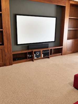 A home theater setup with a large flat-screen TV mounted on a dark wall, surrounded by wooden shelves and cabinets.