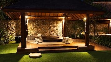 Un área de estar al aire libre con techo, un fondo de pared de piedra, escalones de madera y un arreglo circular de asientos.