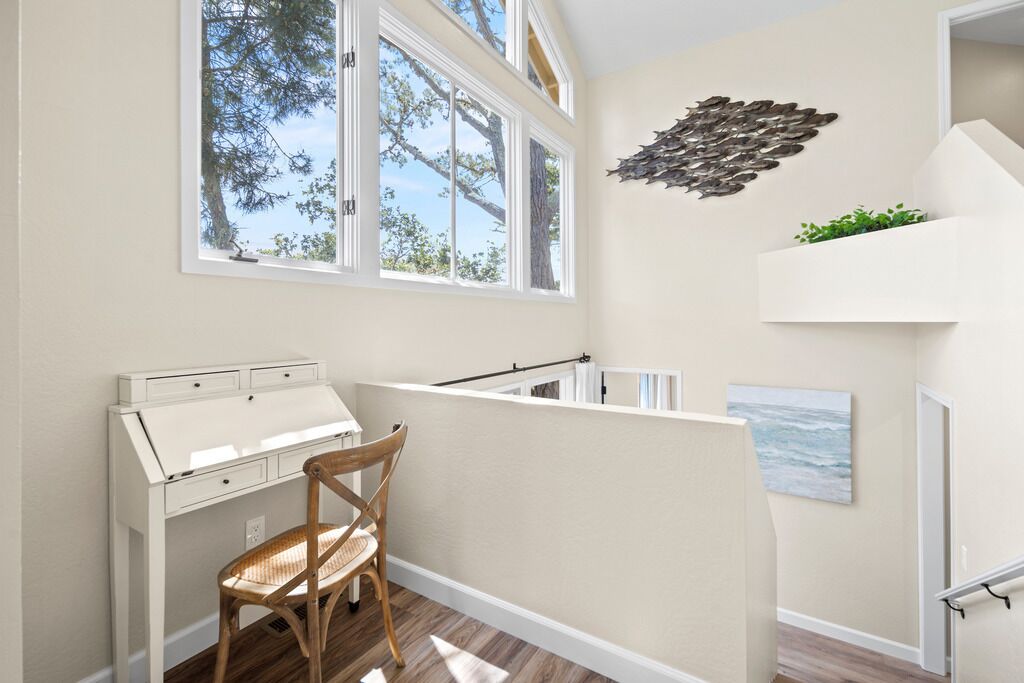 A room with a desk, chair, large windows, a wall art piece, and a view of trees.