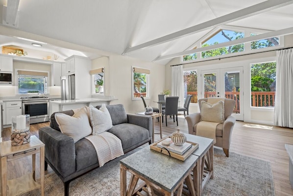 A spacious living area with a grey sofa, a coffee table, and a dining table. There is a kitchen area with white cabinets and a refrigerator. The room has large windows and a view of greenery outside.