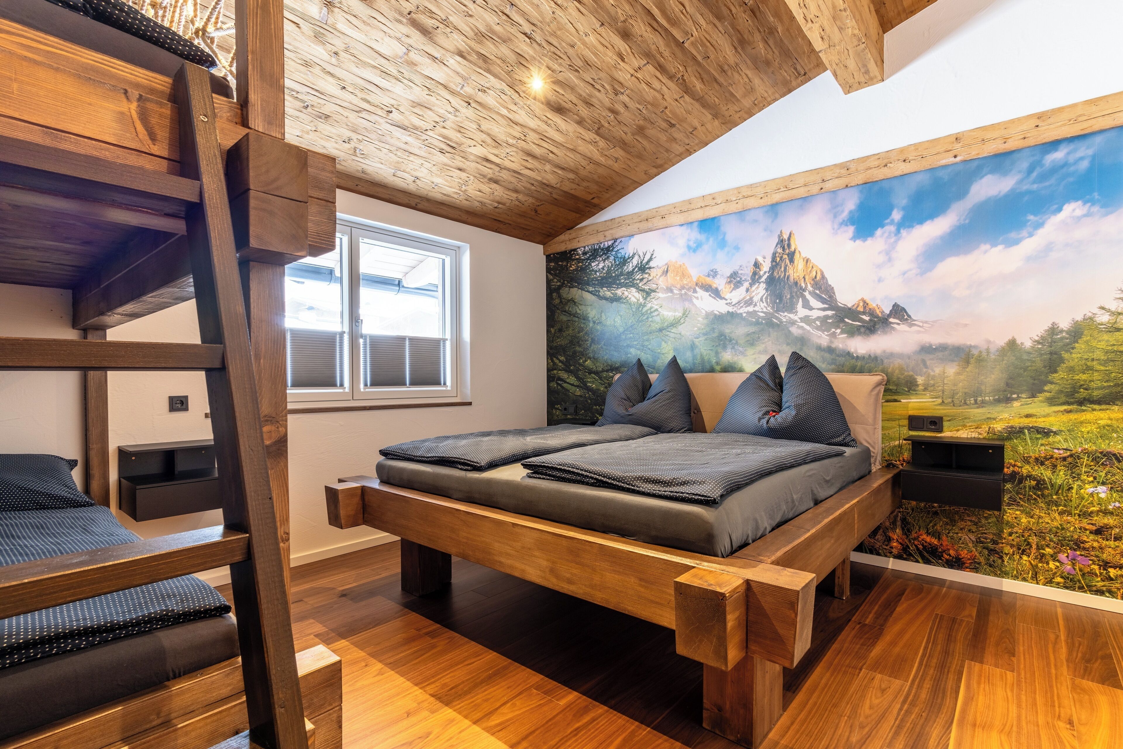 Een slaapkamer met een houten stapelbed, een groot bed met een berglandschap op de muur en een raam.