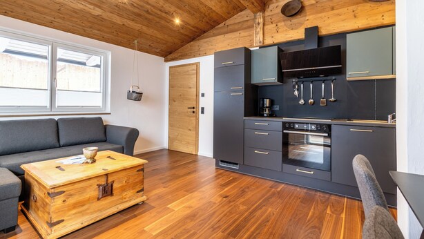 A modern kitchen with wooden ceiling beams, a built-in oven, and a grey sofa in a living area with a wooden chest of drawers. - Appartement Hoamatgfüh (Saalbach)