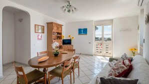 Un coin repas avec une table ronde, des chaises, un téléviseur et un meuble. On y trouve également un canapé avec des coussins et une vue sur l’extérieur grâce à une porte vitrée.