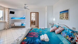 Une chambre à coucher avec un lit recouvert d’une couette aux motifs bleus et rouges évoquant l’océan, un ventilateur de plafond, un téléviseur fixé au mur et une fenêtre avec des rideaux.