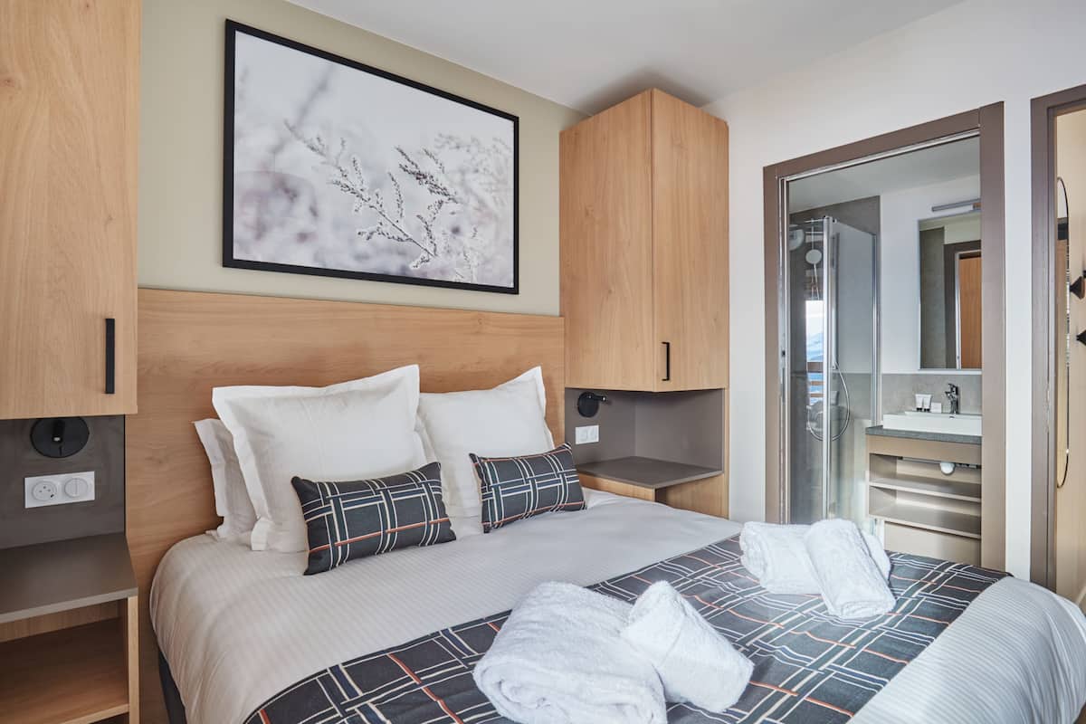 Ein modernes Hotelzimmer mit einem großen Bett, einem hölzernen Kopfteil, einem gerahmten Kunstwerk, einem eingebauten Kleiderschrank und einem Badezimmer mit einer Duschkabine.