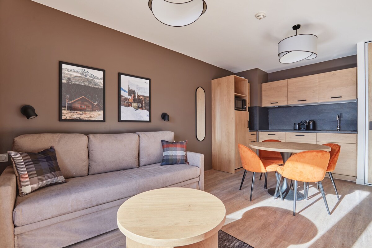 A modern living room with a sofa, a round table, and orange chairs. There is a kitchen area with wooden cabinets and a dining table.