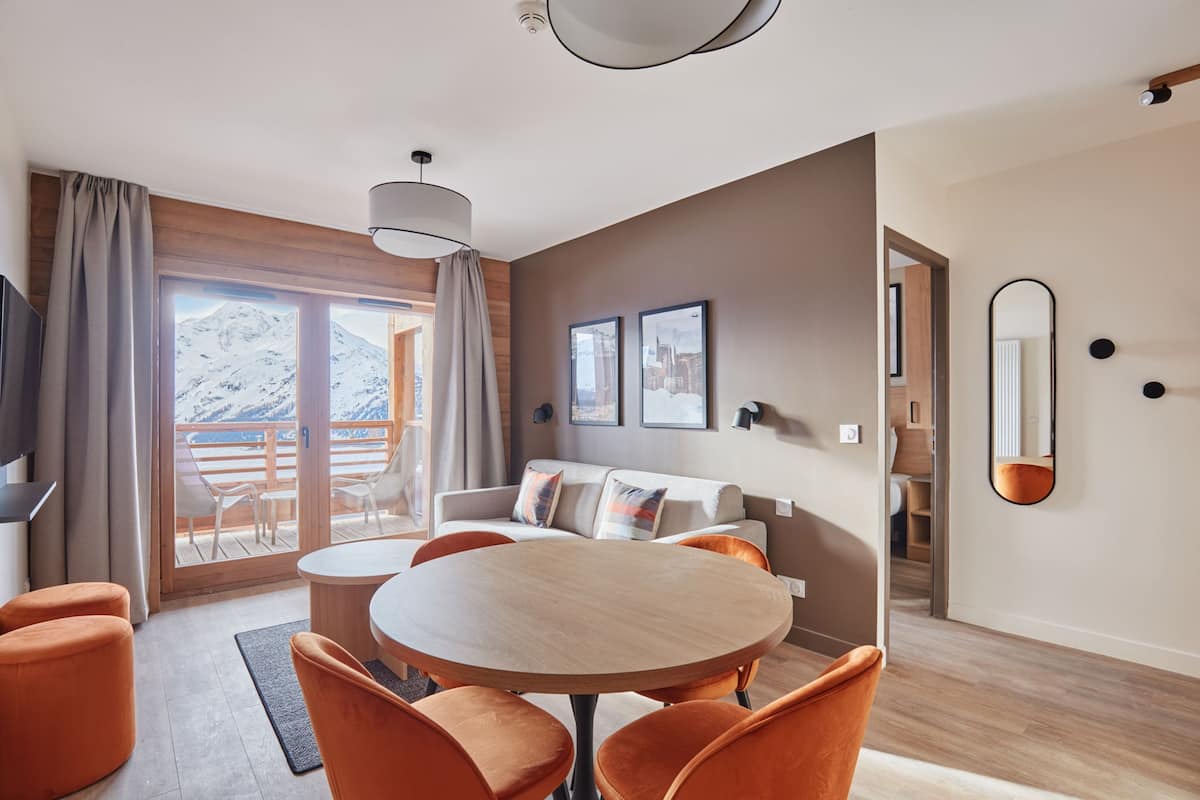 Ein modernes Wohnzimmer mit einem runden Holztisch, orangefarbenen Sesseln, einer Couch mit Kissen, einem Fernseher und einem großen Fenster mit Blick auf schneebedeckte Berge.