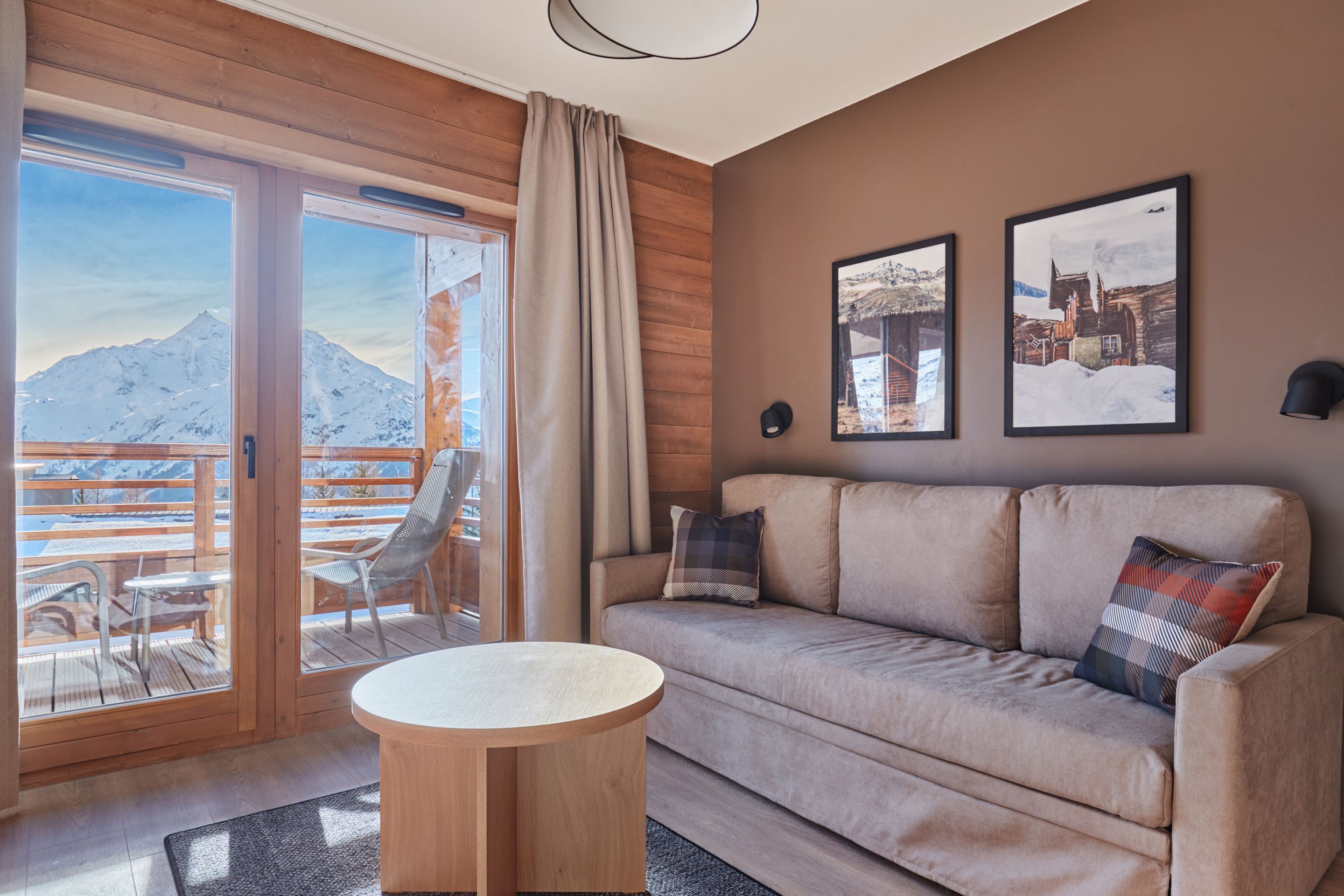 A modern living room with a sofa, a round wooden table, and a balcony with a mountain view.