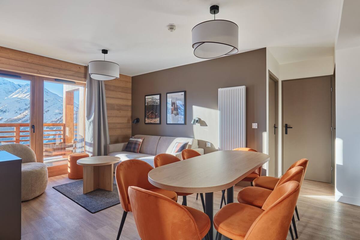 Ein modernes Wohnzimmer mit Holzboden, einem runden Holztisch, orangefarbenen Stühlen, einer Couch mit Kissen und Bergblick durch große Fenster.